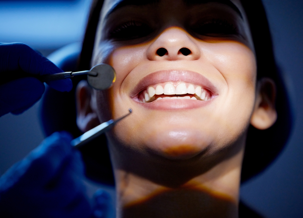 A woman smiles widely during a dental check-up. Gloved hands hold a dental mirror and probe by her teeth. Light and shadow play on her face, revealing her healthy, white teeth clearly.