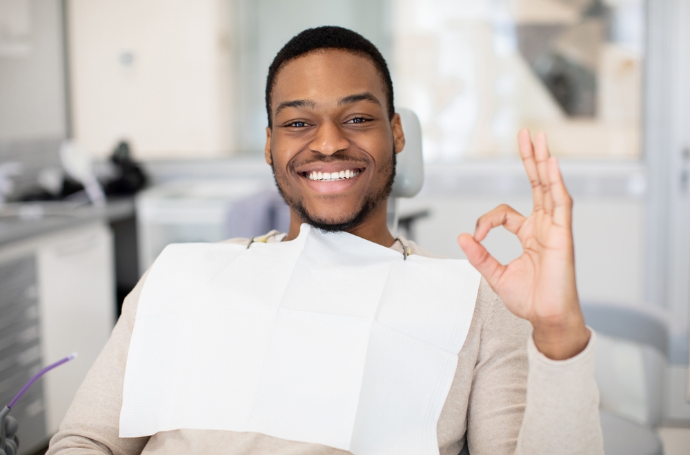 A smiling man sits in a dentist’s chair wearing a dental bib, making an “OK” hand gesture. The dental office background is bright and modern with visible equipment. He looks relaxed and happy.