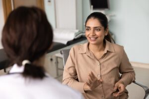 A woman in a beige shirt smiles and gestures as she talks to a healthcare professional wearing a white coat in a medical office, with an exam table visible in the background.